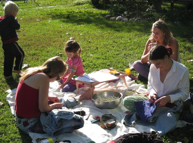 Barn og vaksne på piknikteppe oppå grønt gras. Kvinner syr.