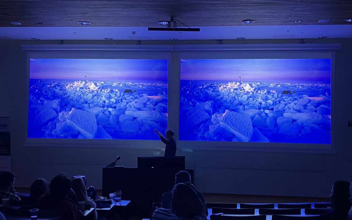 To skjermar i eit auditorium der det står ein mann fremst. Blå, vakre motiv på skjermane frå Arktis (identiske). Mørk sal.