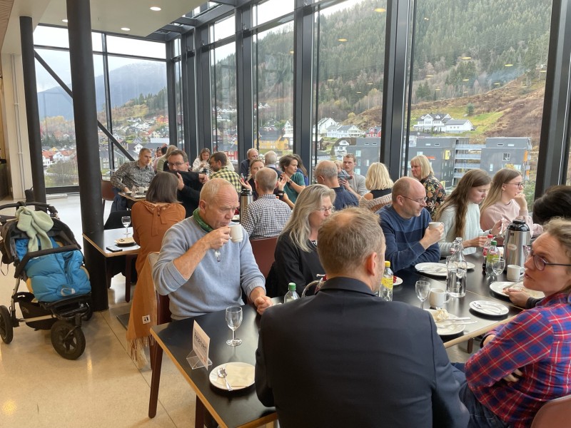 Kantine med tilsette i pent tøy som et lunsj på reserverte bord. Fjellandskap utanfor glasa.