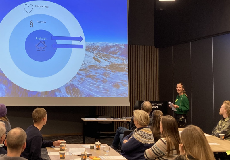 kvinne held fagleg innlegg for menneske plasserte rundt små bord i sal på hotell