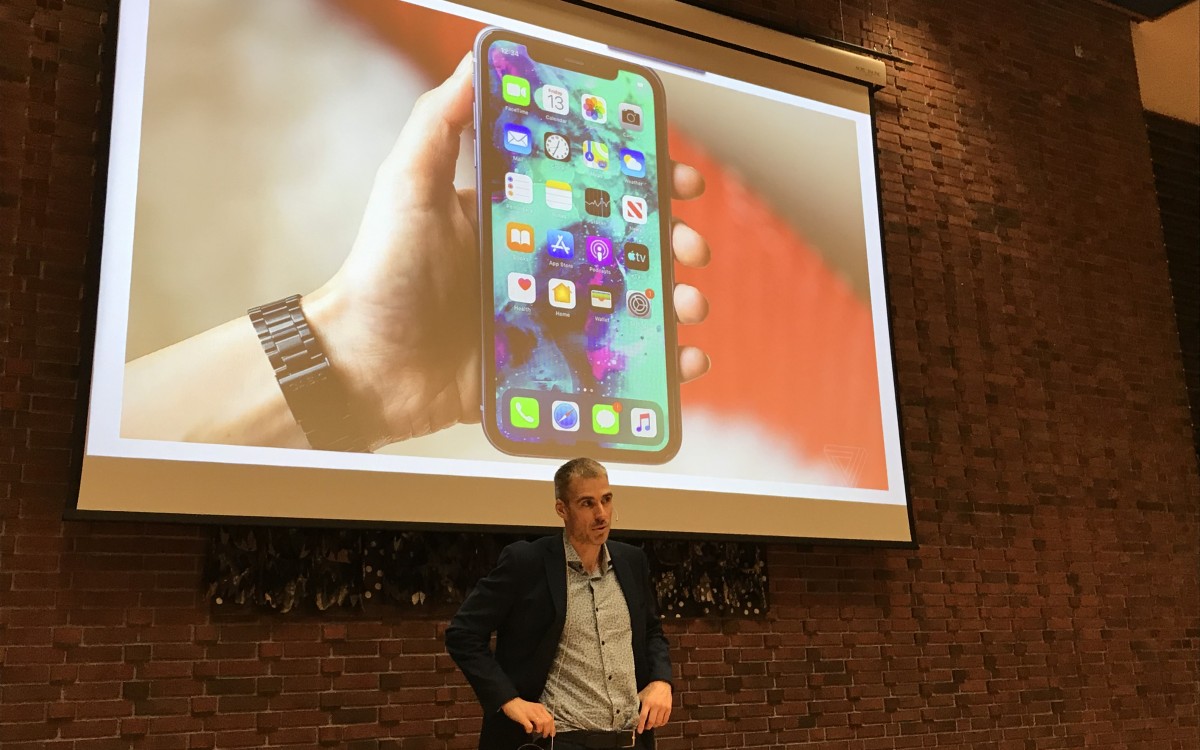 Mann framfor lerret med nærbilde av ei hand som held ein iPhone med stor skjerm.