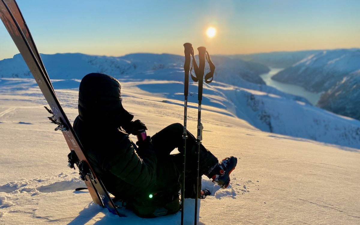 Person med varme kle og hette sit med fjellski og ser ut over vinterfjord frå snølagt fjellplatå. Sol og blå himmel, seint på dag.