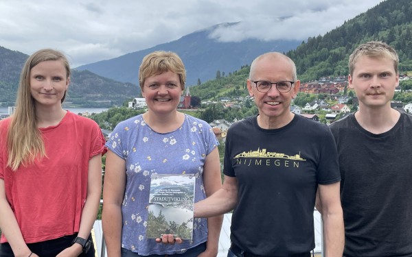 To kvinner og to menn poserer framfor utsikt over Sogndal med bok 