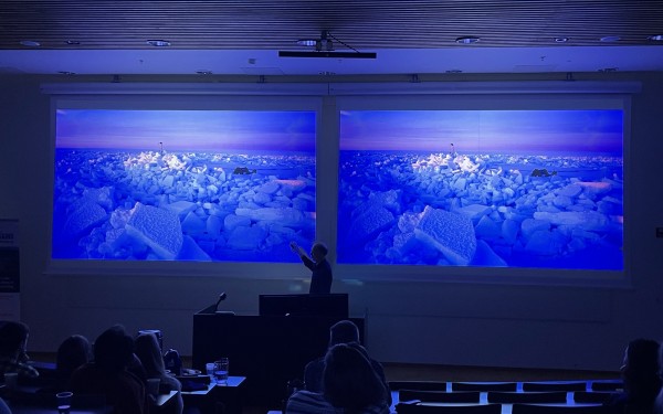 To skjermar i eit auditorium der det står ein mann fremst. Blå, vakre motiv på skjermane frå Arktis (identiske). Mørk sal.