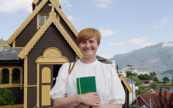 Agnes B. Engeset framfor Engelskkyrkja i Balestrand i kvit bluse og kort, raud frisyre. Blå himmel og lett skydekke. Smiler til kamera. Ho held ei grøn notatbok framfor bringa.