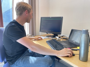 Ung mann i svart t-skjorte og jeans sit ved pult og ser på PC