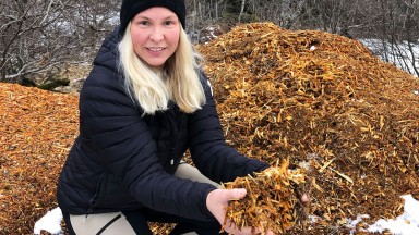 Kvinne i svart jakke og hue sit i ein haug med treflis som ho viser fram i hendene